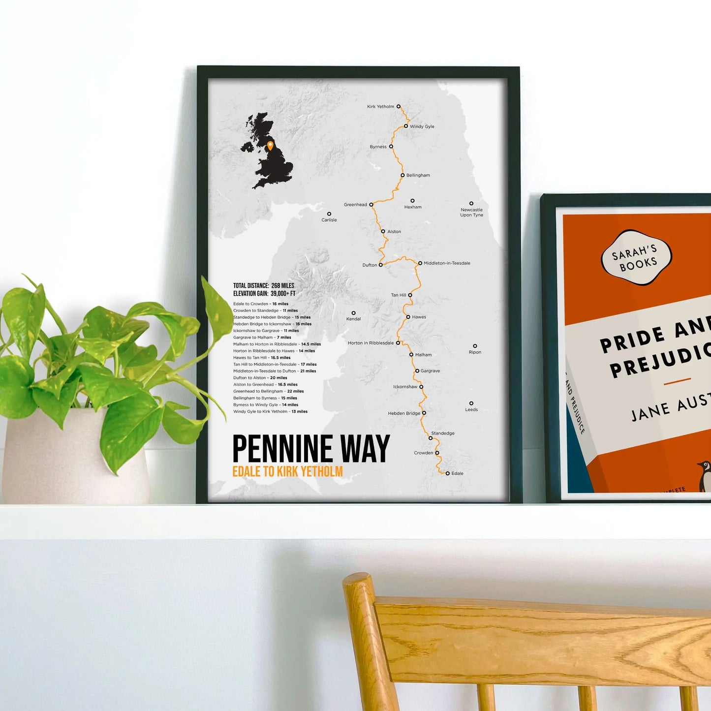 Framed print propped up on a shelf beside a houseplant on the left and another print on the right.