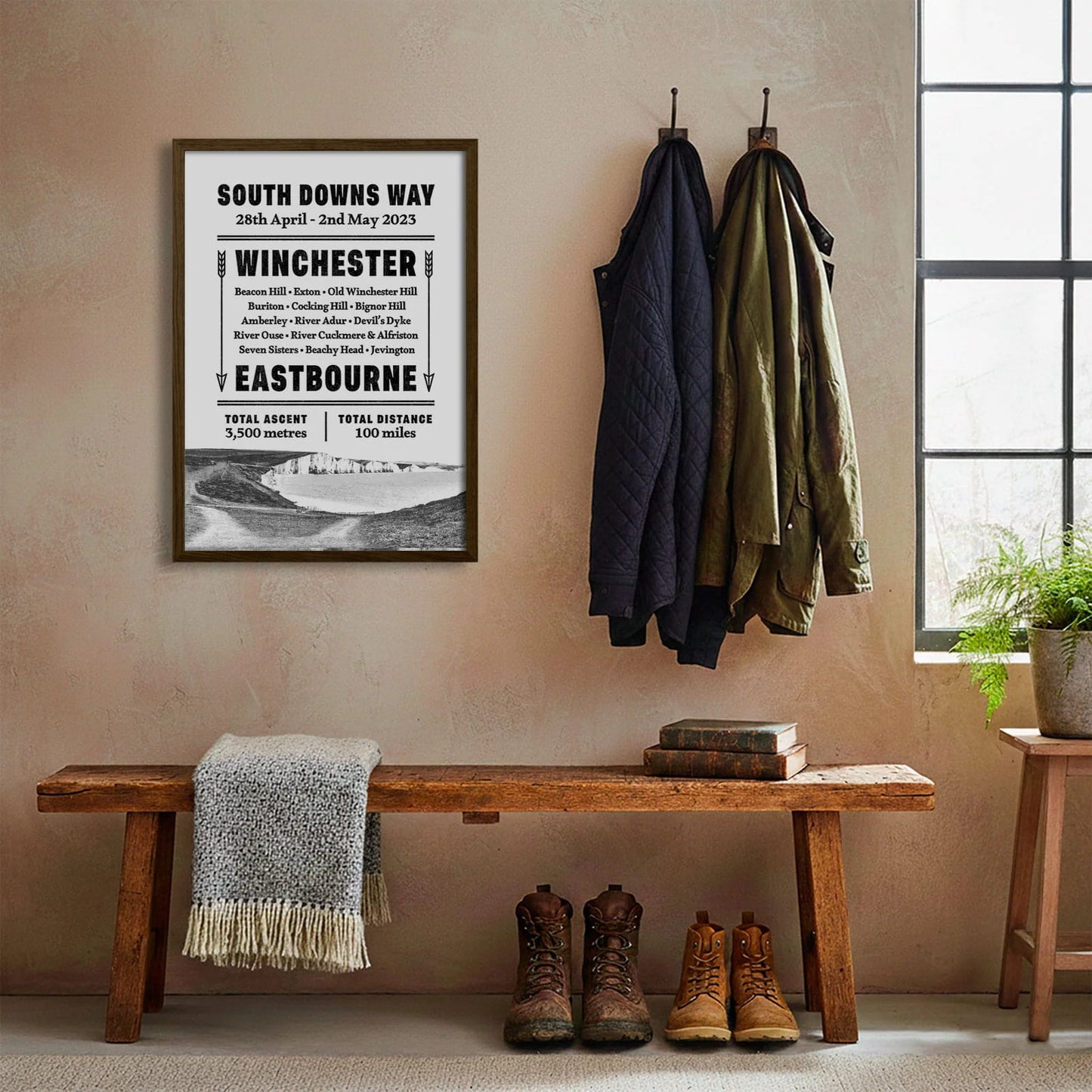 Framed print hanging on cream wall beside coats hanging above bench with walking boots and a scarf