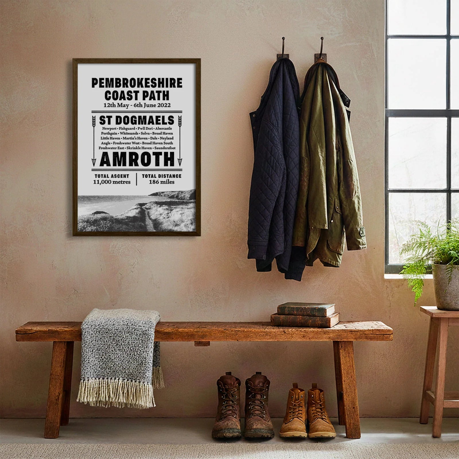 Framed print hanging on cream wall beside coats hanging above bench with walking boots and a scarf