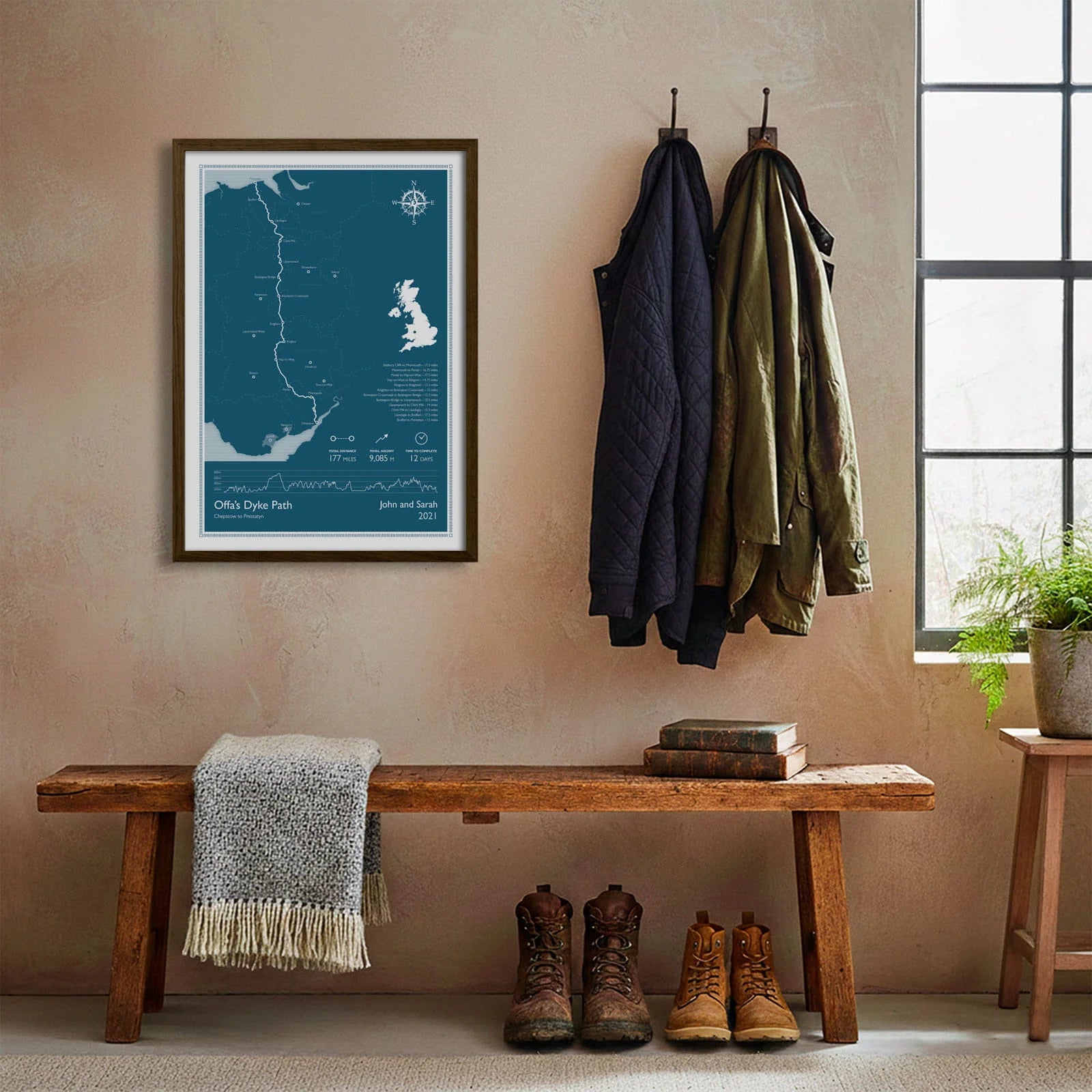 Framed print hanging on cream wall beside coats hanging above bench with walking boots and a scarf