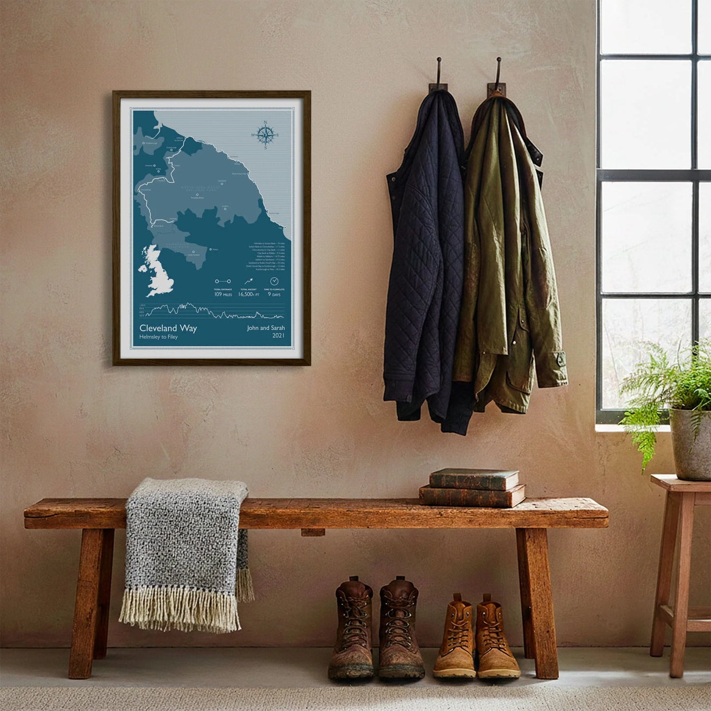 Framed print hanging on cream wall beside coats hanging above bench with walking boots and a scarf