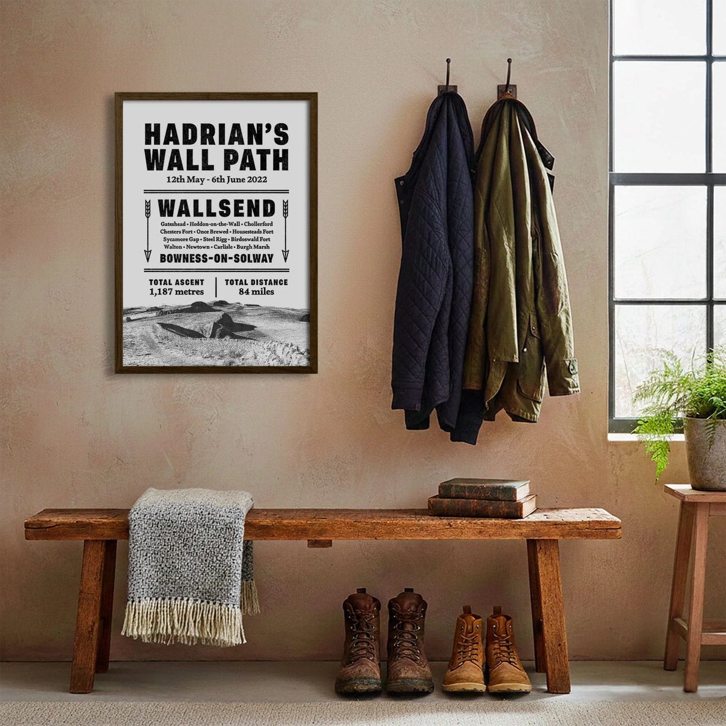 Framed print hanging on cream wall beside coats hanging above bench with walking boots and a scarf