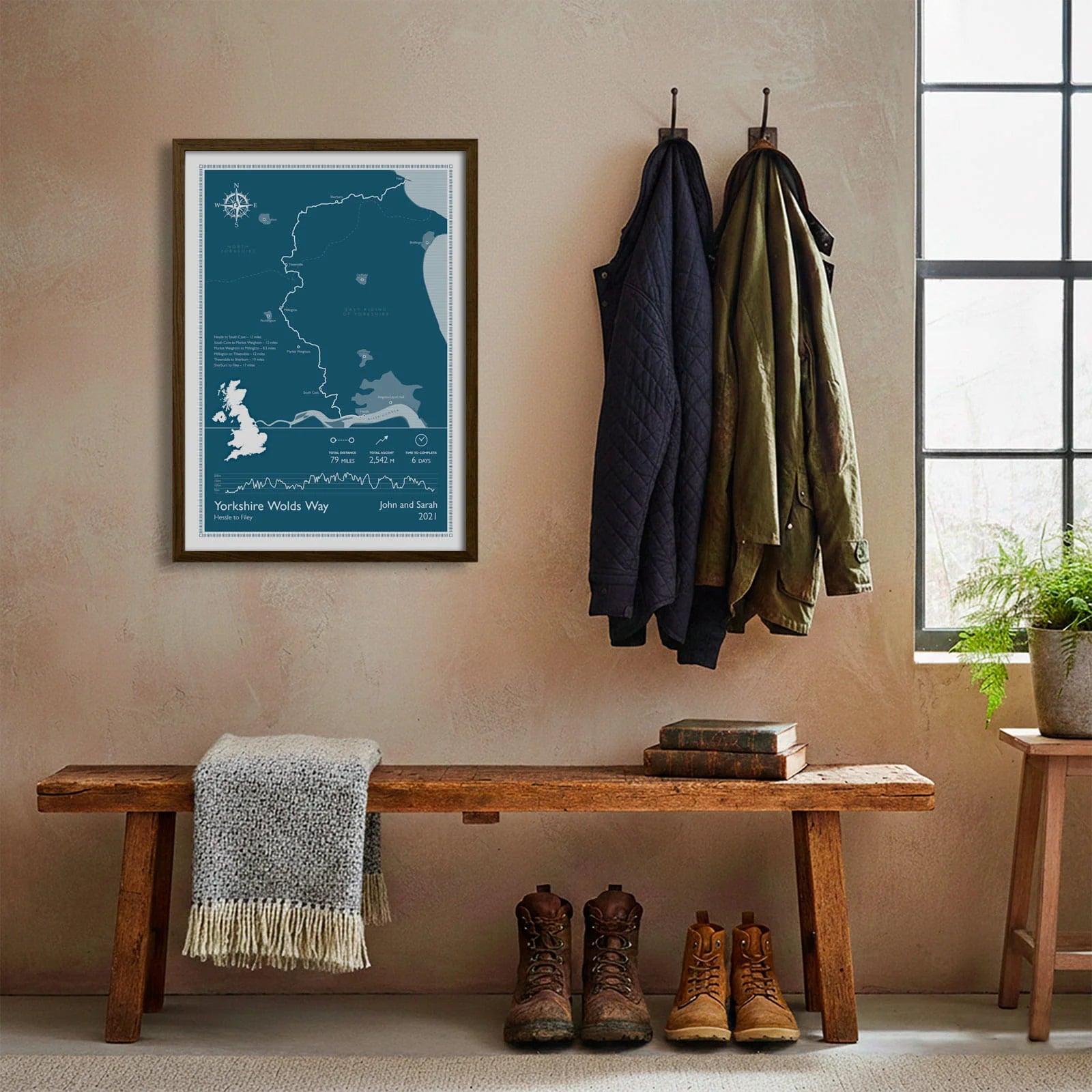 Framed print hanging on cream wall beside coats hanging above bench with walking boots and a scarf