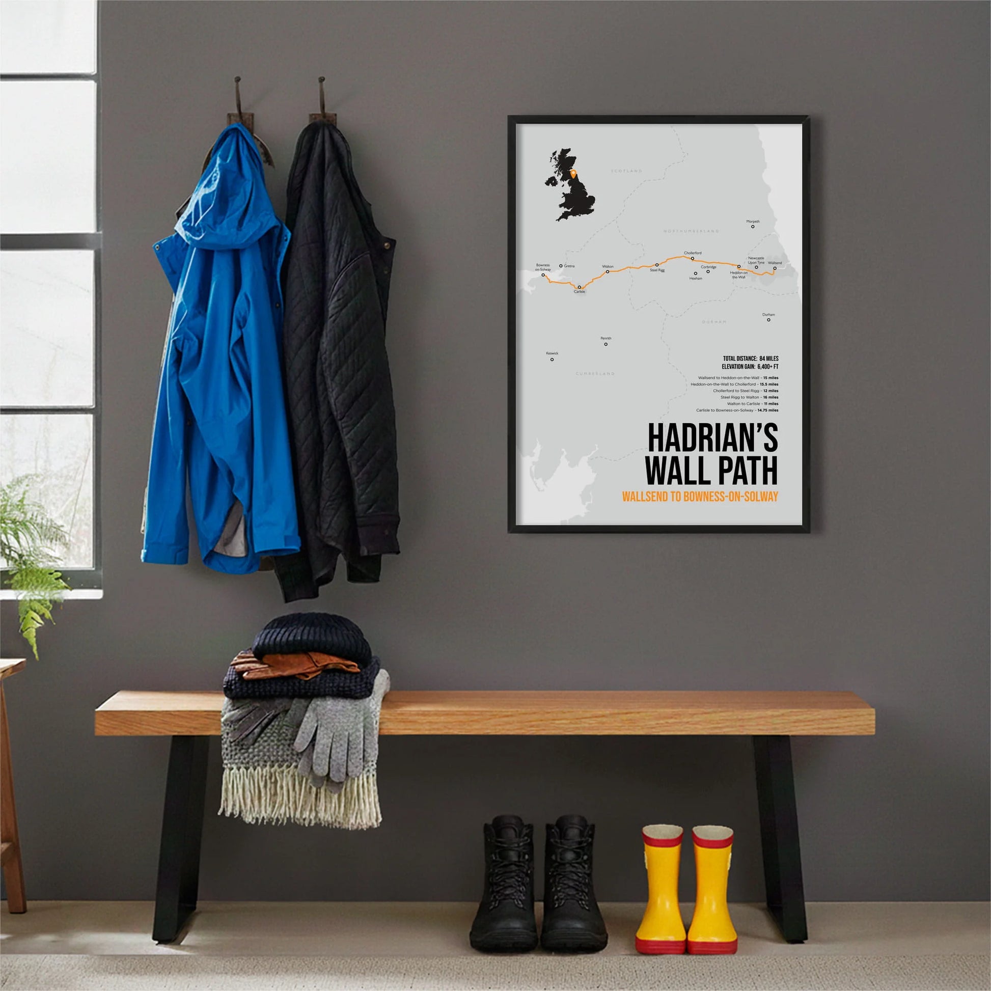 Framed print hanging on cream wall beside coats hanging above bench with walking boots and a scarf