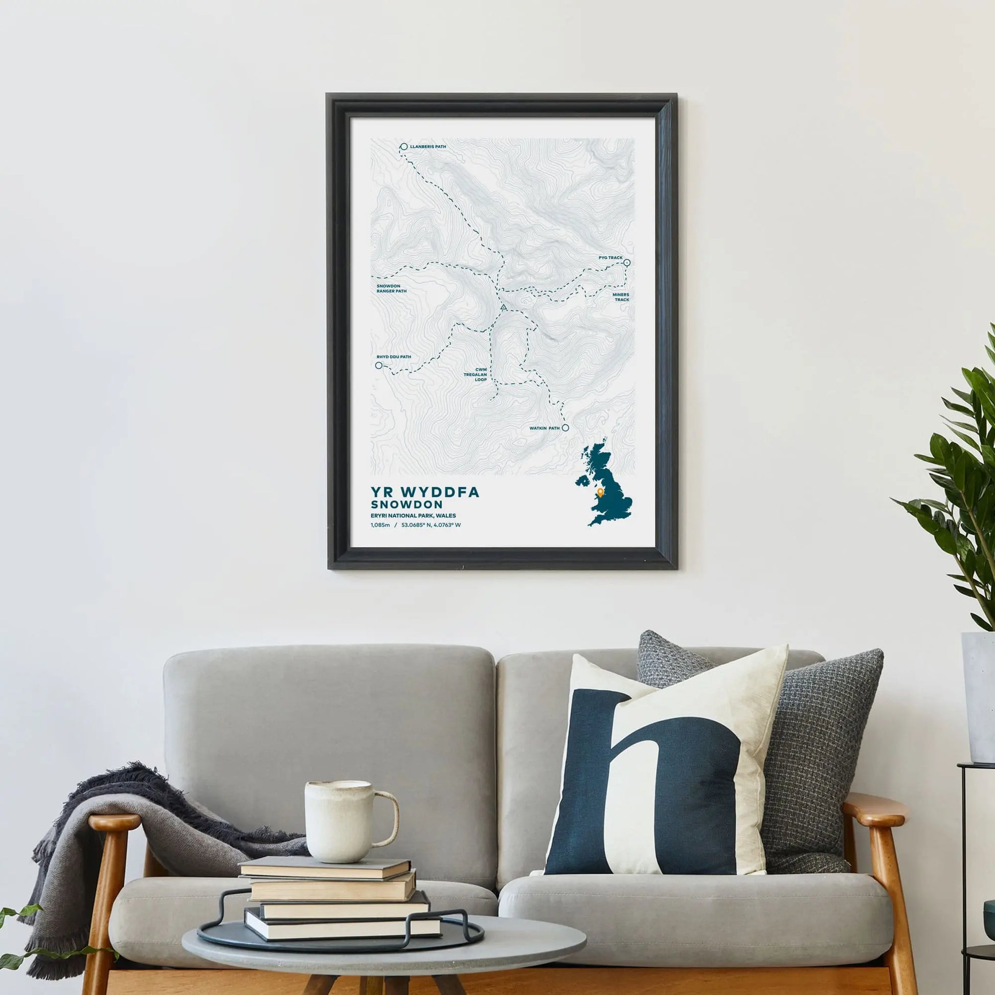 Large print on wall behind a 2 seater sofa covered in cushions with a coffee table covered in books.