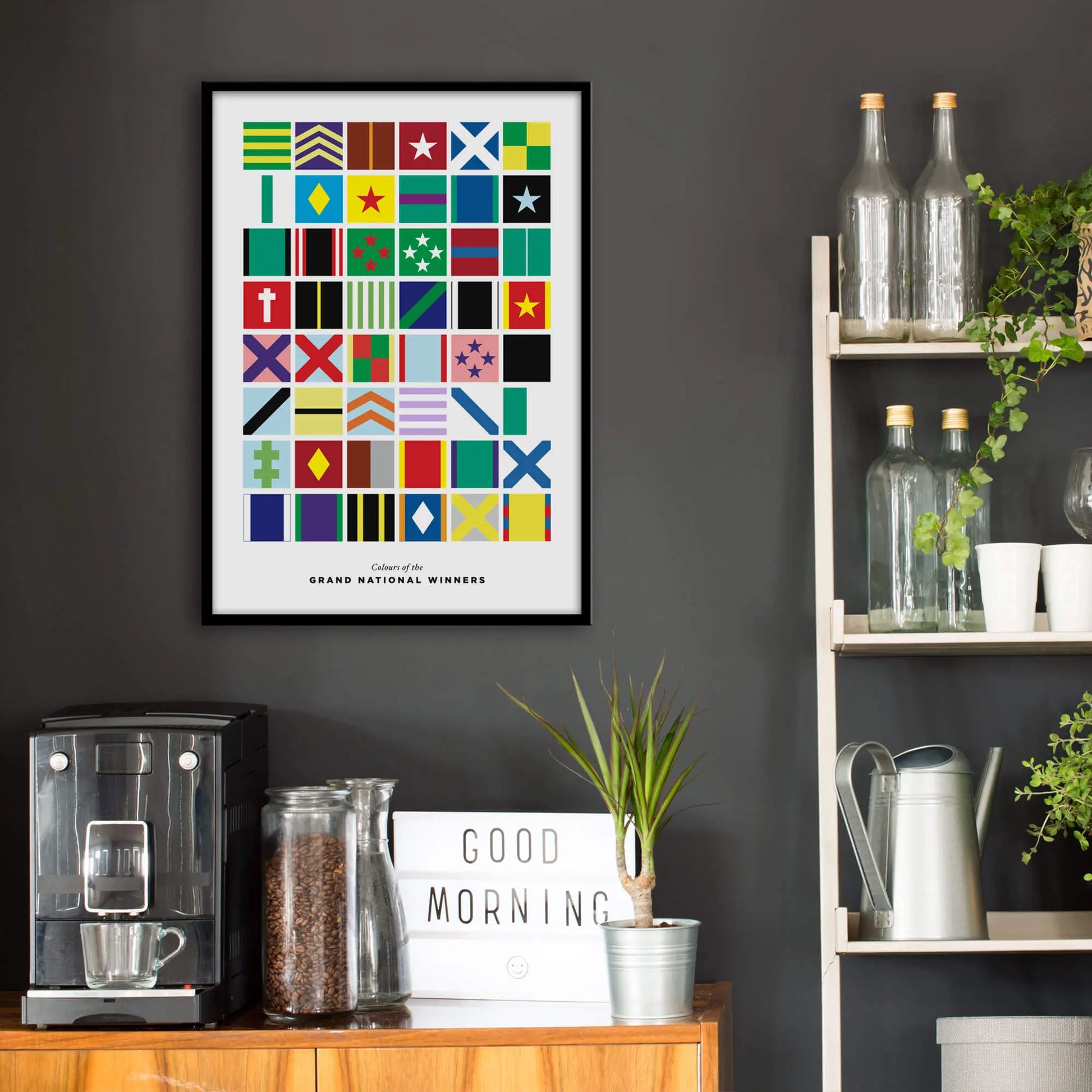 Kitchen wall infront of which is a sideboard with a coffee machine and a set of shelves holding bottles and plants. On the wall above the sideboard is the framed print in a black frame on a dark grey wall.